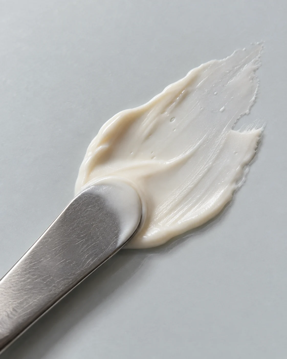 Cream texture study: an irregular ivory smear and a brushed silver cosmetic spatula