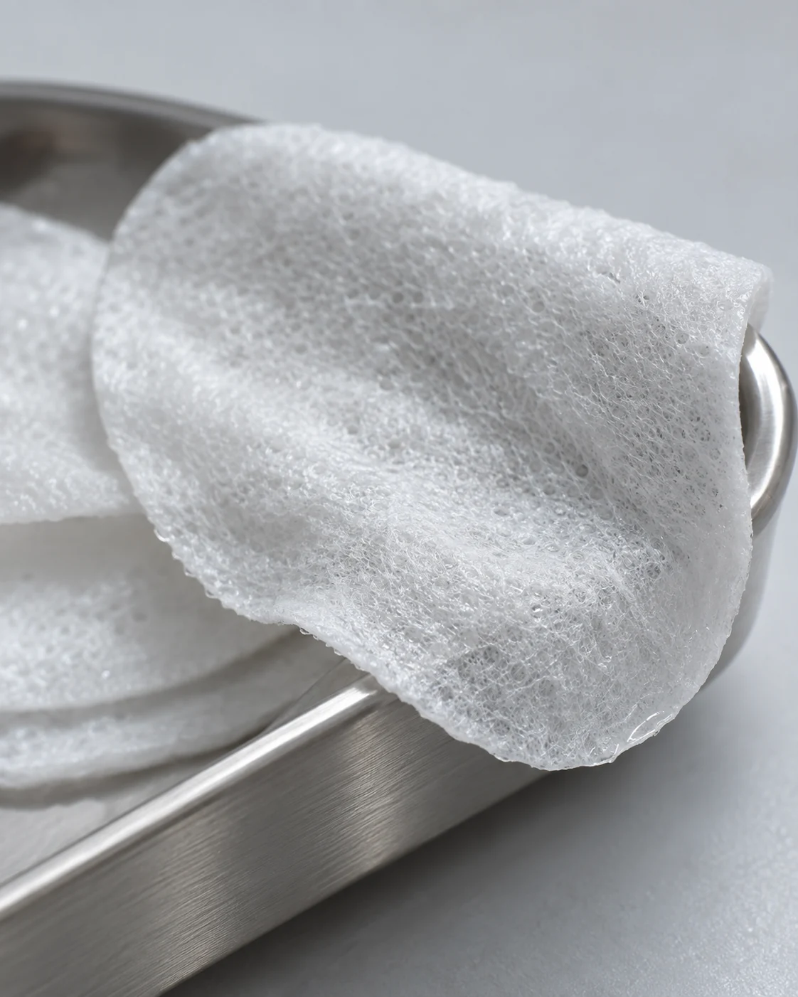 Close material study of a thin damp exfoliating pad showing fine fibers over a brushed silver tray
