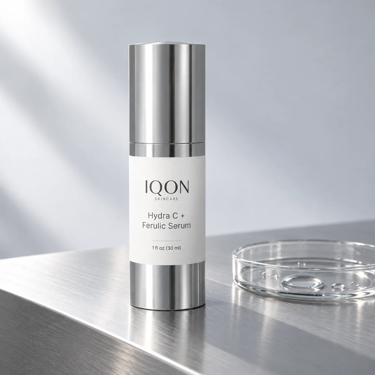 IQON Hydra C + Ferulic Serum on brushed silver beside a clear glass dish