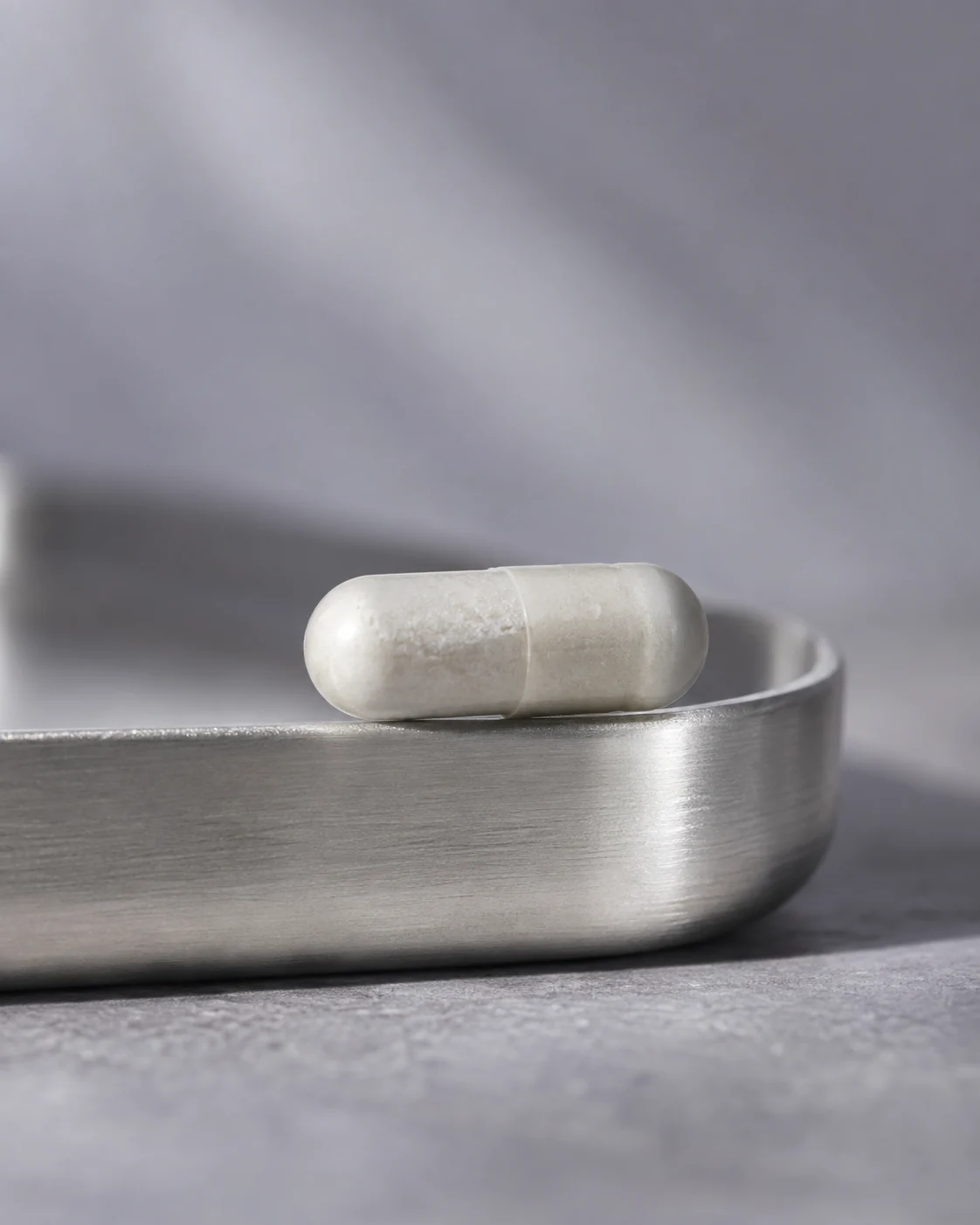 One white capsule on the edge of a brushed silver tray against a muted lavender-gray background