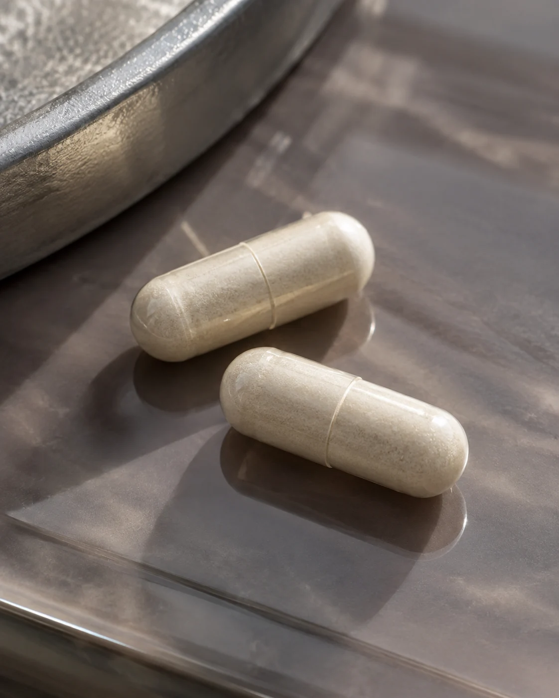 Two ivory capsules resting diagonally on smoky mauve-gray glass in soft directional light